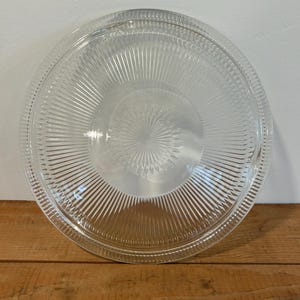 Pode incluir: Um prato de bolo de vidro transparente com uma borda canelada e um centro elevado. O prato é projetado para segurar um bolo ou outra sobremesa.