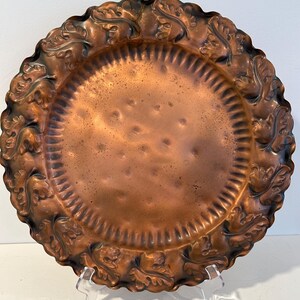May include: A large, round copper serving tray with a scalloped edge and a raised, embossed design of leaves around the rim. The tray has a hammered finish and a warm, aged patina.