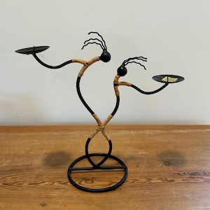 May include: Black metal candle holder with two figures, each holding a small, round tray. The figures have black heads and arms, with woven tan material around their bodies. The base is a black, circular design.
