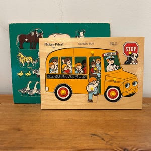 May include: A wooden Fisher-Price School Bus puzzle with 13 pieces, featuring a yellow bus, a driver, and children. The board displays the text 'SCHOOL BUS', 'AGES 3-6', and '13 PIECES'. Behind it is a green wooden puzzle board showing various farm animals.