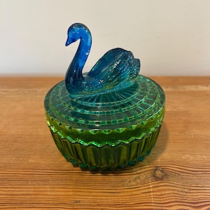 May include: A vintage, teal and green glass trinket box with a swan lid. The swan is a deep blue color, and the box has a geometric design. The box is sitting on a wooden surface.