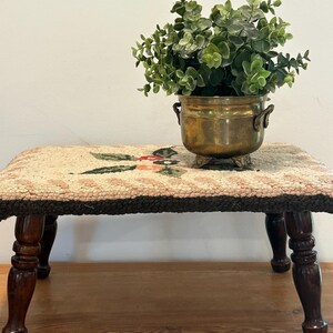 May include: A small, antique footstool with a floral needlepoint top and dark wooden legs. A brass pot with green foliage sits on top of the footstool, adding a touch of vintage charm. The footstool measures approximately 12 inches long, 8 inches wide, and 10 inches tall.