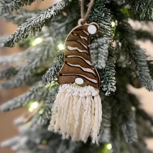 May include: A wooden gnome ornament with a brown and white striped hat and a white yarn beard. The ornament is hanging on a Christmas tree.