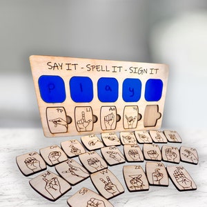 May include: Wooden educational toy with the text "SAY IT - SPELL IT - SIGN IT". The board has blue letter slots spelling "PLAY" and corresponding sign language illustrations. Additional wooden tiles with sign language symbols are included.
