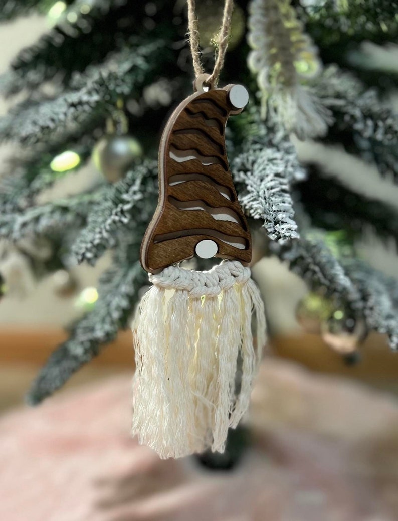 May include: A wooden gnome ornament with a white beard made of yarn. The gnome is wearing a brown hat with white stripes and has a white pom-pom on top.