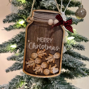 May include: A wooden Christmas ornament shaped like a mason jar with the words "Merry Christmas" written on it. The jar is filled with gingerbread people with names written on them.