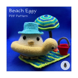 May include: A crocheted yellow chick wearing a blue hat and a yellow inflatable ring, sitting on a green and yellow striped beach towel with a red bucket and a green and blue striped umbrella.