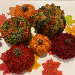 以下が含まれることがあります： 緑、オレンジ、ワインレッドなど、さまざまな色と質感のクロシェ編みかぼちゃが5つあります。かぼちゃは、秋の色の葉が敷き詰められた白い表面に配置されています。