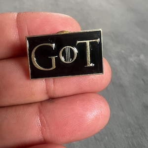 May include: A black enamel pin with gold lettering that reads "GOT".