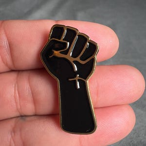 Peut inclure: Poing noir en émail levé dans les airs avec un contour doré. Le poing est un symbole de force et de solidarité.