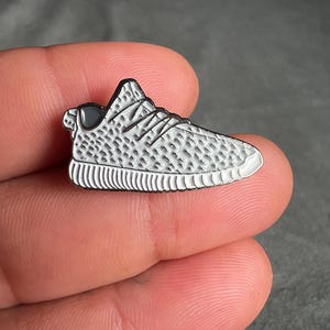 May include: A silver-colored enamel pin shaped like a white sneaker with black accents. The pin has a textured design and is a popular fashion accessory.
