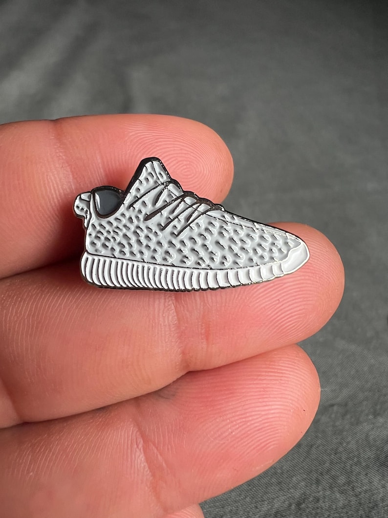 May include: A silver-colored enamel pin shaped like a white sneaker with black accents. The pin has a textured design and is a popular fashion accessory.