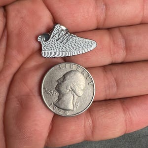 May include: A silver-colored pin of a white sneaker with a textured design. The pin is about the size of a US quarter.