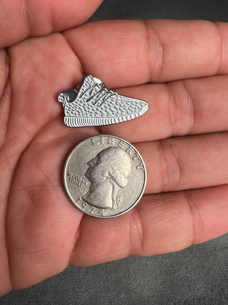 May include: A silver-colored pin of a white sneaker with a textured design. The pin is about the size of a US quarter.