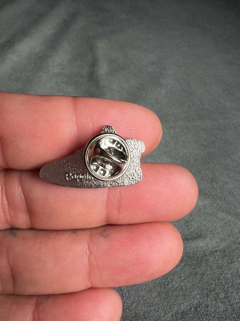 May include: Silver-toned metal pin with a black outline and a silver-toned pin back. The pin is shaped like a shoe and has the word "Darth" printed on the back.