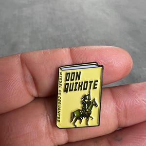 Puede incluir: Un pin de esmalte amarillo con forma de libro con el título "DON QUIJOTE" y el autor "MIGUEL DE CERVANTES". El pin presenta una ilustración de dibujos animados de Don Quijote montando un caballo.