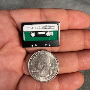 May include: A black and green cassette tape pin with the words "Good Vibes" written on the top. The pin is about the size of a US quarter.
