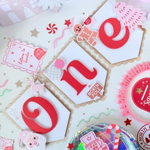 May include: A festive birthday banner with the word "one" in red, surrounded by Christmas-themed decorations. Includes a gingerbread man, candy canes, and a plate with a nutcracker design. The color palette is primarily red, white, and pink.