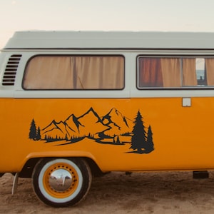 May include: A black mountain and forest decal is applied to the side of a yellow and white vintage van. The decal features a mountain range, trees, and a river, creating a nature-themed design.