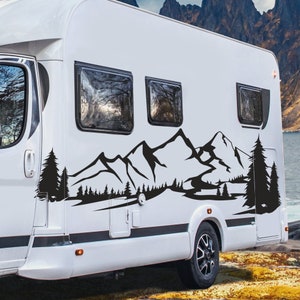 May include: A white recreational vehicle with a black mountain and forest decal on the side. The decal features a silhouette of a mountain range and pine trees. The RV has three windows and a black tire.