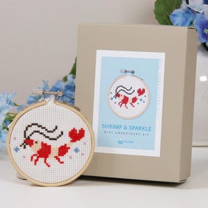 Può includere: Un mini kit da ricamo con un disegno a punto croce di un gambero rosso con antenne nere e accenti a forma di cuore. Il kit include un telaio in legno e una scatola con il testo "SHRIMP & SPARKLE MINI EMBROIDERY KIT".