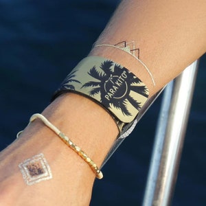 May include: A gold and black wristband with a palm tree design and the text "PARA KITO" on it. The wristband is worn on a person's wrist.