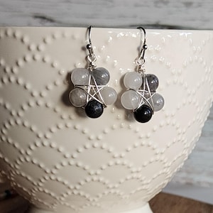 May include: A pair of silver wire wrapped earrings with a five-pointed star design. The earrings feature grey and black gemstone beads.