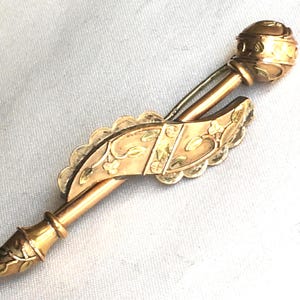 May include: A gold-toned antique brooch with intricate floral and geometric designs. The brooch features a decorative pin and a detailed, rounded end. The piece has a scalloped edge and a detailed, rounded end.