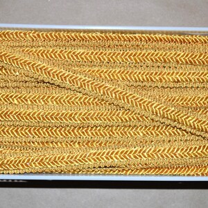 May include: A spool of gold-colored gimp trim featuring a consistent herringbone or chevron weave pattern. The metallic-toned decorative cord is wound tightly around a white plastic reel, displaying a continuous braided texture throughout the length.