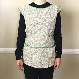 May include: A vintage-style apron with a floral pattern in pastel colors. The apron has a white base with star and flower designs in shades of yellow, blue, and purple. It features green trim along the edges and pockets, and a fitted waist.