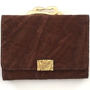May include: A brown suede wallet with a gold-colored clasp and decorative gold-colored accents. The wallet has a textured surface and a rectangular shape. The clasp is a unique design, adding to the wallet's aesthetic appeal.