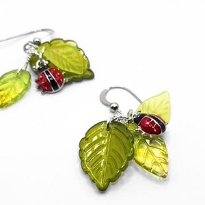 May include: A pair of dangle earrings featuring green leaf charms and red ladybug accents. The earrings have silver-colored hooks and are set against a white background. The leaves are a translucent green and yellow.