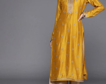 Puede incluir: Una kurta de manga larga de color amarillo mostaza con bordados dorados y un pantalón palazzo beige a juego. La kurta tiene un escote redondo y una abertura lateral. La modelo lleva joyas doradas y sandalias.