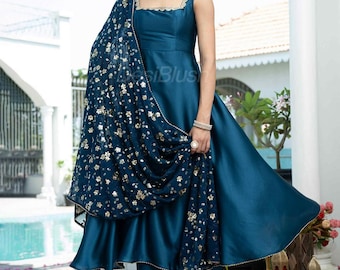 Puede incluir: Un atuendo formal de color verde azulado con un vestido sin mangas con cuello cuadrado y pantalones a juego. Un chal estampado con detalles dorados cae sobre los hombros. El conjunto se complementa con joyas y sandalias doradas.