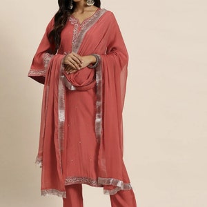 May include: A coral pink salwar kameez with silver embroidery and a matching dupatta. The outfit features a V-neckline, long sleeves, and a straight-cut kameez. The dupatta has a silver border and fringe.