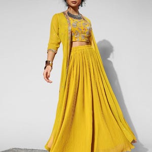 May include: A mustard yellow three-piece outfit featuring a crop top, pleated skirt, and long jacket. The crop top has silver embroidery. The outfit is part of the DesiBlush collection.