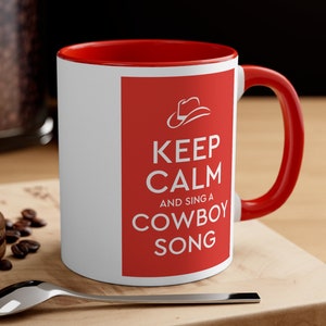 Op de afbeelding: Witte keramische mok met een rode rand en handvat. De mok heeft een rode achtergrond met witte tekst die "Keep Calm and Sing a Cowboy Song" luidt. Boven de tekst staat een wit cowboyhoed-icoon.
