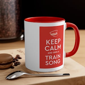 Op de afbeelding: Een witte keramische mok met een rode rand en handvat. De mok heeft een rood vierkant met een witte treinconducteurpet en de tekst "Keep Calm and Sing a Train Song".