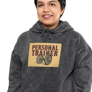 Könnte beinhalten: Ein schwarzer Kapuzenpullover mit Used-Look. Die Vorderseite des Pullovers zeigt eine Grafik im Vintage-Stil mit dem Text "PERSONAL TRAINER" und einem Bild eines Gewichts.