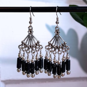 May include: A pair of silver-coloured dangle earrings. Each earring has a fan-shaped top with dangling strands of silver beads and black cylindrical beads. The earrings are suspended from silver fish hook ear wires.