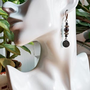 May include: A single dangle earring with a bronze-colored hook and a series of beads. The beads include dark red, black, and clear crystal accents. A small, round charm with a Celtic knot design hangs at the bottom.