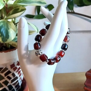 May include: A stretch bracelet featuring polished, deep red, square-shaped beads interspersed with small, round, silver-toned spacer beads and one larger, spherical, silver-toned bead with a detailed, open-work, cut-out pattern design.