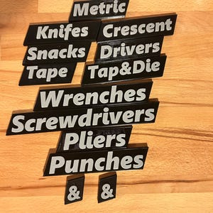 May include: Black and white labels with tool and item names such as "Metric", "Knives", "Snacks", "Tape", "Crescent", "Drivers", "Tap & Die", "Wrenches", "Screwdrivers", "Pliers", and "Punches". The labels are on a wooden surface.