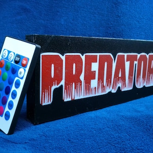May include: A black wooden box with the word "PREDATOR" in red letters. The box is sitting on a blue surface. A remote control with a variety of colored buttons is next to the box.