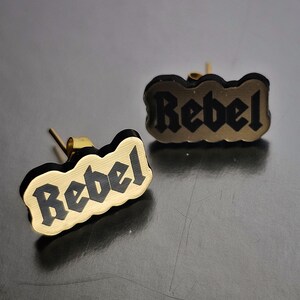May include: A pair of gold-colored stud earrings. Each earring features the word "Rebel" in black, gothic-style lettering, set against a gold background. The earrings have a unique, irregular shape.