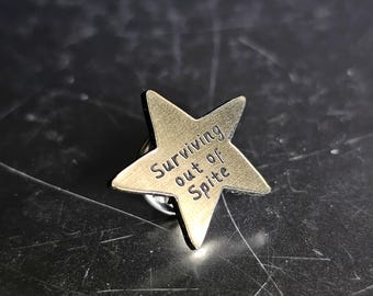May include: A bronze-colored star-shaped pin with the phrase "Surviving out of Spite" engraved on its surface. The pin has a classic star design with five points and a pin backing for attachment to clothing or accessories.