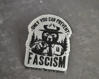 May include: A silver-colored metal pin featuring a bear in a ranger hat, pointing. The text reads "ONLY YOU CAN PREVENT FASCISM" in a circular pattern and "FASCISM" below the bear.