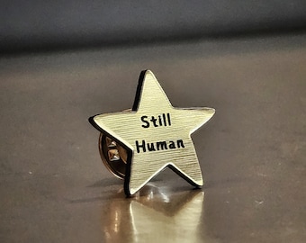 May include: A gold-colored star-shaped pin with the words "Still Human" engraved in black. The pin has a classic design and a secure clasp on the back. The pin is displayed on a reflective surface.