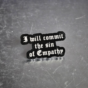 May include: A black pin with the phrase "I will commit the sin of Empathy" in white gothic lettering. The pin has a rounded, irregular shape and is set against a metallic surface.