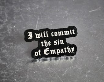 May include: A black pin with the phrase "I will commit the sin of Empathy" in white gothic lettering. The pin has a rounded, irregular shape and is set against a metallic surface.